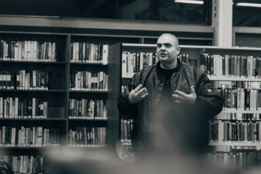 Matt Vapor speaking to a seated audience in a library, gesturing with both hands, tall bookshelves behind him. Black and white photograph.