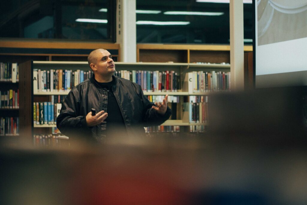 Matt Vapor presenting in a library, holding a remote and looking toward a projection screen, with bookshelves behind him.