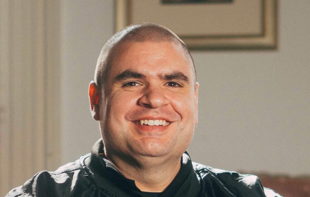 Matt Vapor with a shaved head wearing a black leather jacket, sitting indoors with a beige wall and framed picture behind him.