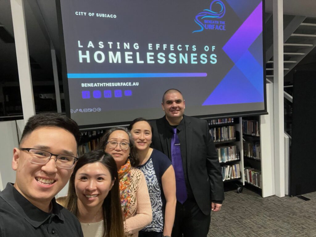 Five people smiling together for a group photograph in a library, in front of a screen showing the City of Subiaco 'Lasting Effects of Homelessness' title slide.