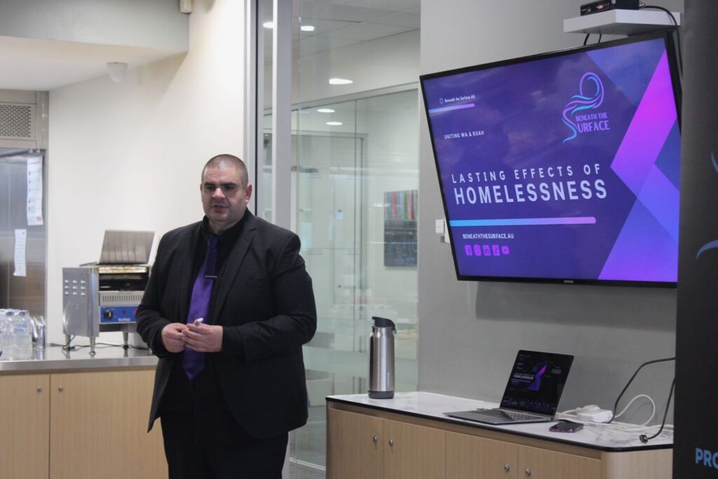 Matt Vapor in a dark suit and purple tie standing beside a wall-mounted screen displaying the 'Lasting Effects of Homelessness' title slide, with a laptop set up on a bench beside him.