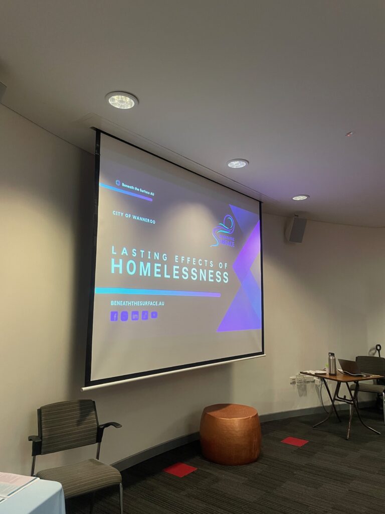 A projector screen in an empty meeting room showing the City of Wanneroo 'Lasting Effects of Homelessness' title slide, with a chair and side table nearby.