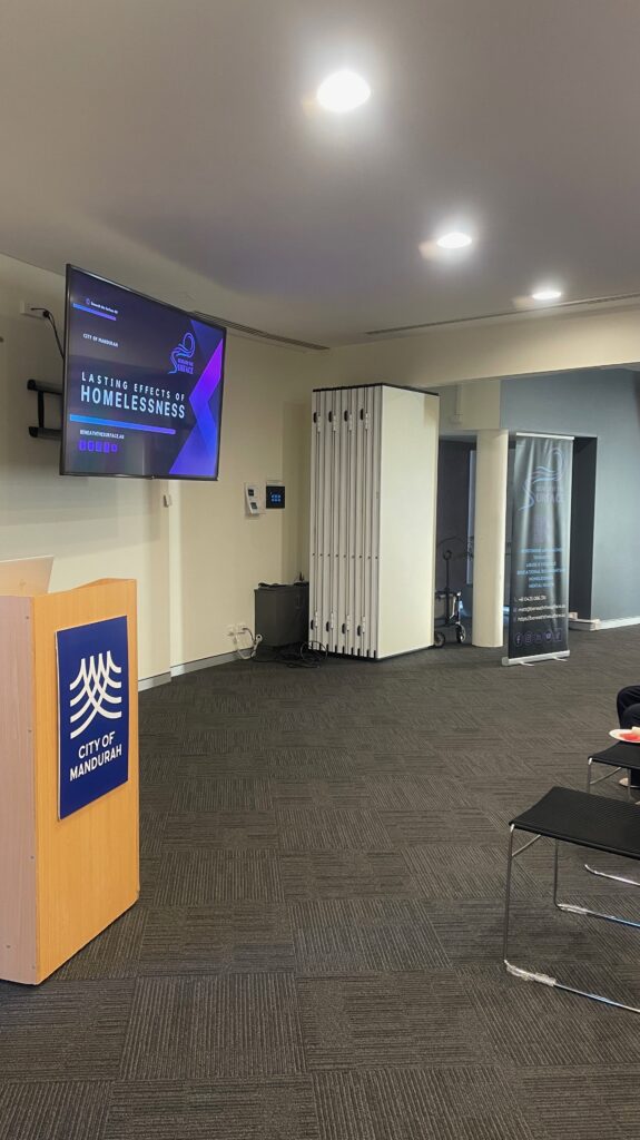 A meeting room set for a presentation, with a wall-mounted screen showing the 'Lasting Effects of Homelessness' title slide and a City of Mandurah lectern in the foreground.
