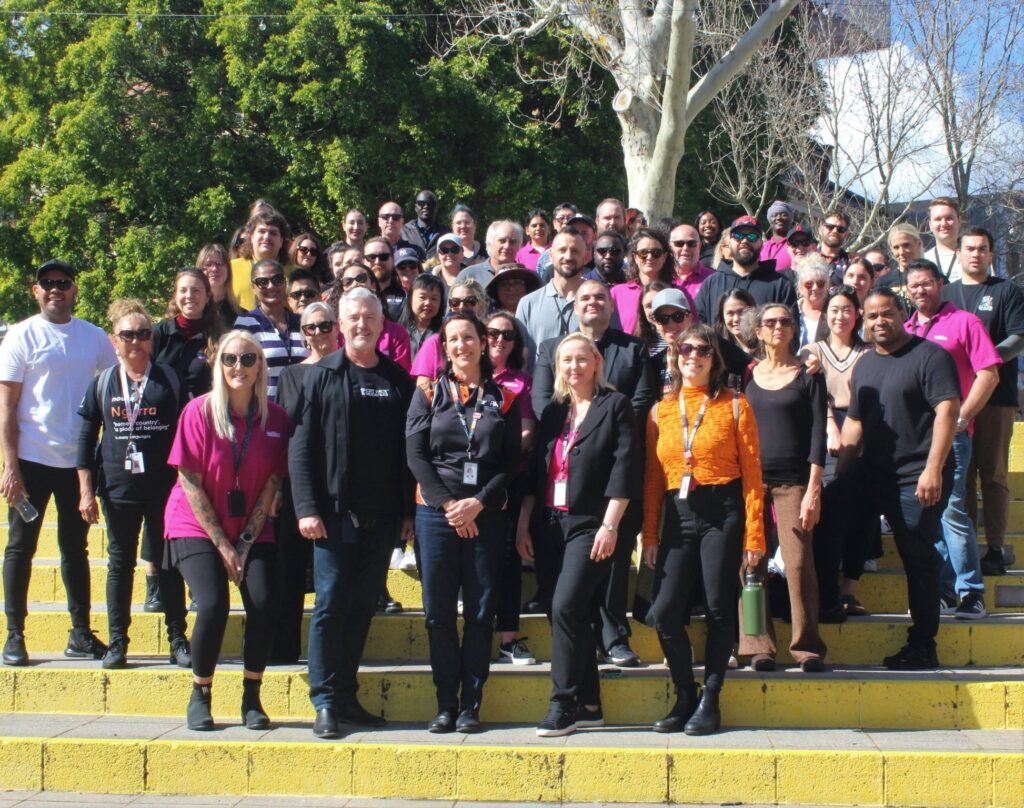 A large group of roughly fifty people posed together on outdoor steps in bright sunshine, many wearing pink shirts and lanyards.