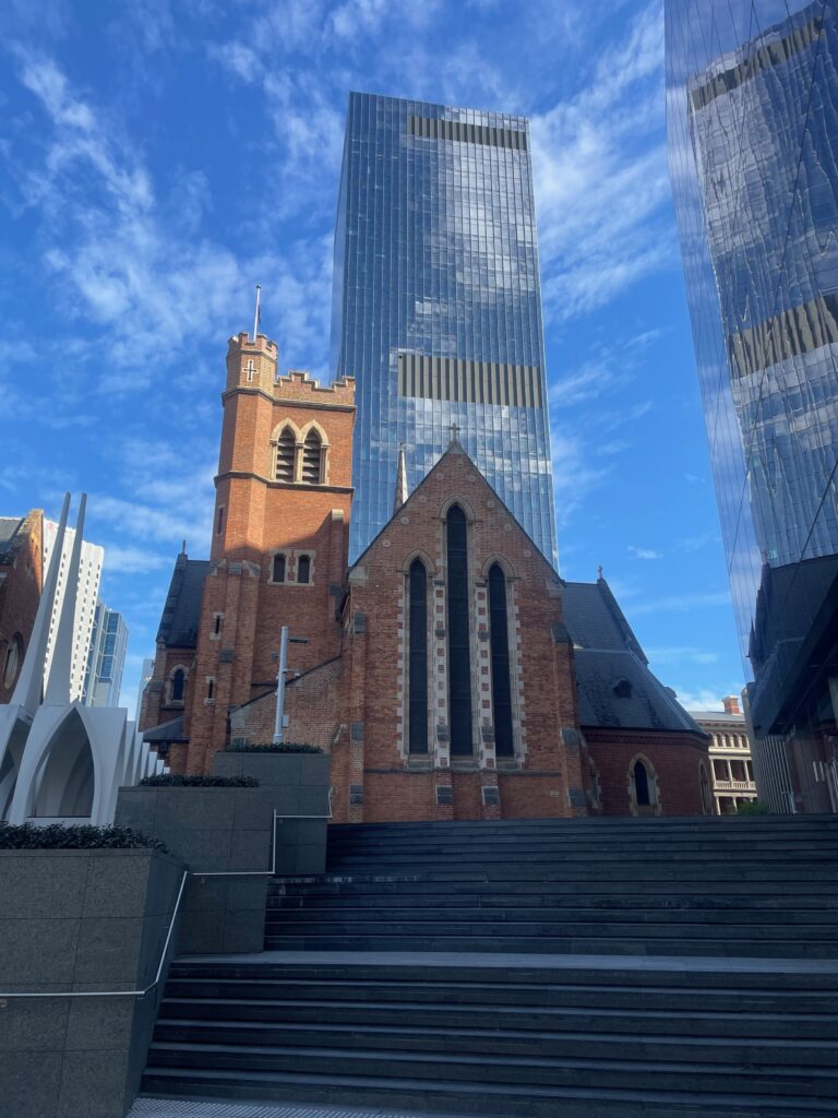 A red brick Gothic-style church with a square bell tower, framed against a modern glass office tower under a blue sky, with wide stone steps in the foreground.