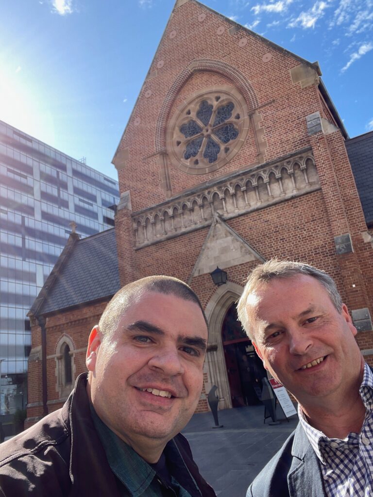Matt Vapor and another man smiling for a selfie outside a red brick church with a rose window above its arched entrance.
