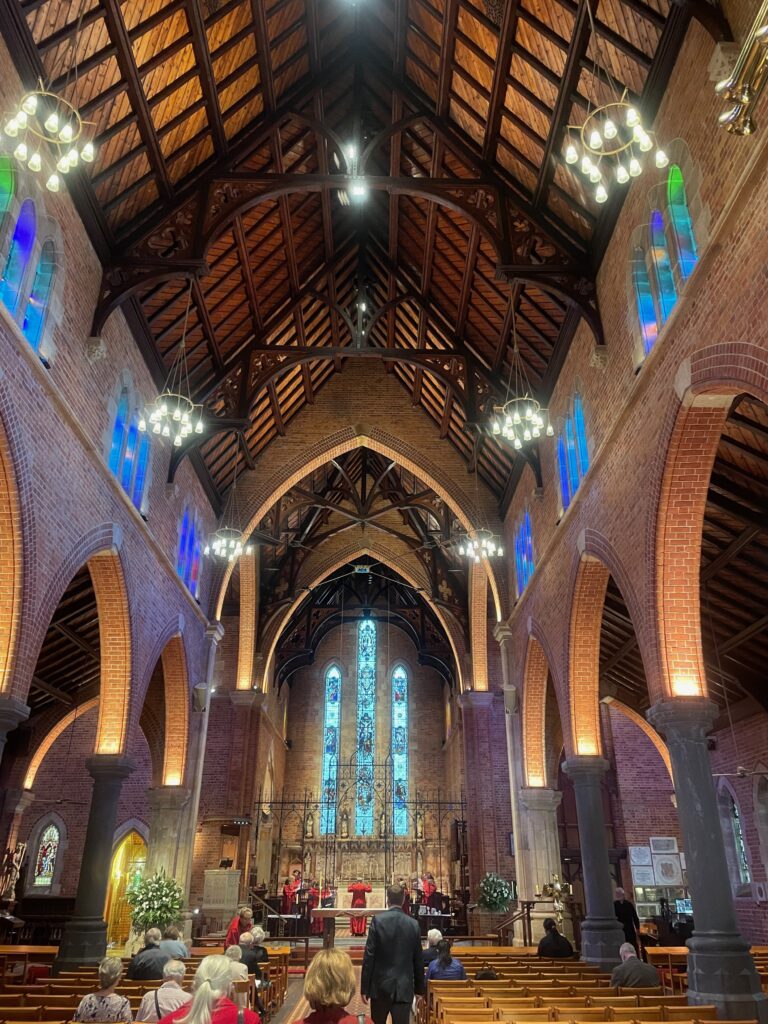 The interior of a brick church during a service, looking down the nave toward the altar where clergy in red vestments are gathered, with the congregation seated in wooden pews.