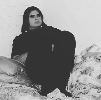 Matt Vapor as a young man with long dark hair, sitting hunched against a bare brick wall on a mattress with scattered bedding, knees drawn up to his chest. Black and white photograph.