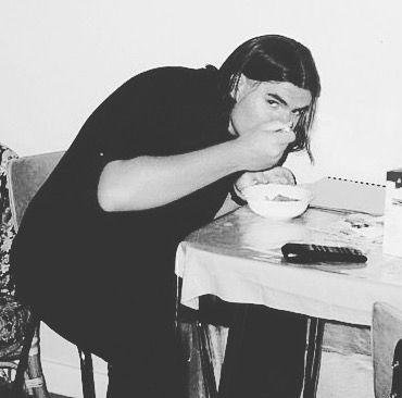 Matt Vapor as a young man with long dark hair, leaning over a small table and eating from a bowl. Black and white photograph.