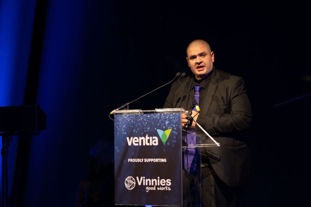 Matt Vapor speaking from a lectern on a darkened stage, wearing a dark suit and purple tie. The lectern reads: Ventia, proudly supporting Vinnies Good Works.